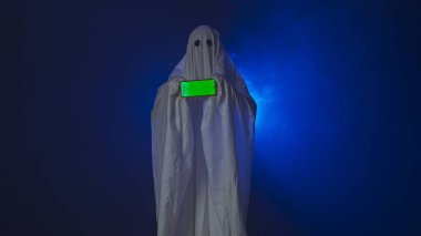 Kameraya poz veren hayalet gibi giyinmiş biri. Beyaz çarşaflı komik cadılar bayramı hayaleti stüdyodaki koyu mavi arka planda akıllı telefon kroma anahtar yeşil ekran gösteriyor. Mutlu Cadılar Bayramı ürkütücü konsepti.