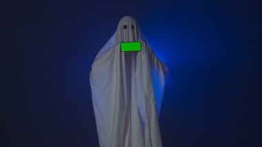 Kameraya poz veren hayalet gibi giyinmiş biri. Beyaz çarşaflı komik cadılar bayramı hayaleti stüdyodaki koyu mavi arka planda akıllı telefon kroma anahtar yeşil ekran gösteriyor. Mutlu Cadılar Bayramı ürkütücü konsepti.