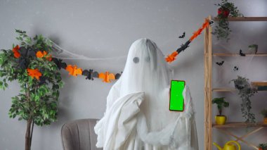 Kameraya poz veren hayalet gibi giyinmiş biri. Beyaz çarşaflı komik cadılar bayramı hayaleti parlak odada siyah gözlü akıllı telefon kromu yeşil ekranını işaret ediyor. Mutlu Cadılar Bayramı ürkütücü konsepti.