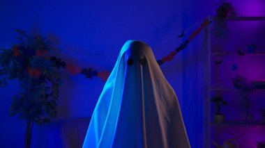 Kameraya poz veren hayalet gibi giyinmiş biri. Karanlık odada siyah gözlü beyaz çarşaflı komik bir cadılar bayramı hayaleti ve çarşafın altında bir fener yaktı. Mutlu Cadılar Bayramı ürkütücü konsepti.