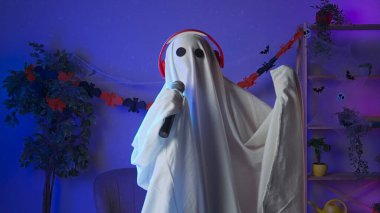 Kameraya poz veren hayalet gibi giyinmiş biri. Beyaz çarşaflı komik cadılar bayramı hayaleti. Karanlık odada siyah gözleri var. Kulaklıkla müzik eşliğinde karaoke mikrofonunda şarkı söylüyor. Mutlu Cadılar Bayramı ürkütücü konsepti.