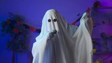 Kameraya poz veren hayalet gibi giyinmiş biri. Karanlık odada siyah gözlü, beyaz çarşaflı komik cadılar bayramı hayaleti müzik eşliğinde dans ediyor ve karaoke mikrofonunda şarkı söylüyor. Mutlu Cadılar Bayramı ürkütücü konsepti.
