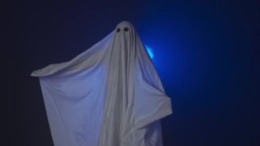 Kameraya poz veren hayalet gibi giyinmiş biri. Beyaz çarşaflı komik cadılar bayramı hayaleti arka planda korkunç jestler yapıyor ve stüdyoda mavi spot ışıkları var. Mutlu Cadılar Bayramı ürkütücü konsepti.