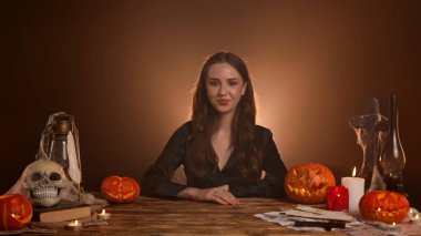 Büyücülük yapan mistik kadın büyücü. Cadılar Bayramı gotik cadı falcı masasında oturmuş, mutlu bir gülümsemeyle parmaklarıyla işaret ediyor. Sihirli cadılar bayramı kavramı.