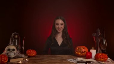 Büyücülük yapan mistik kadın büyücü. Cadılar Bayramı gotik cadı falcısı masa başında oturmuş, yan boşluğu işaret ediyor, gülümseyen göz kırpıyor ve kabulle başını sallıyor. Sihirli cadılar bayramı kavramı.