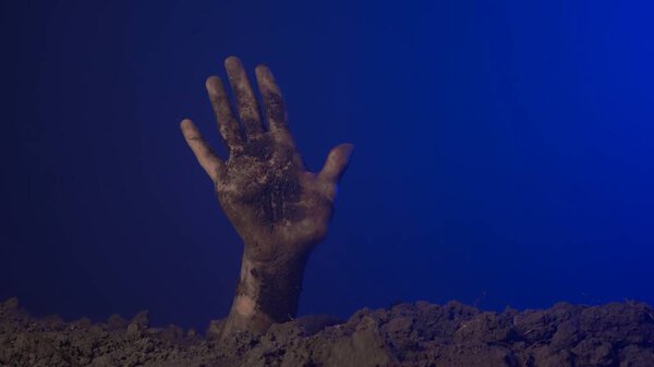 Creepy monster hand shows gestures in graveyard. Scary dirty halloween zombie hand raising up from grave ground shows high five open palm, isolated on blue background. Mystery halloween concept.