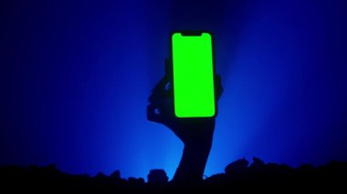 Mezarlıkta ürkütücü canavar hareketler yapıyor. Mezardan yükselen korkunç pis cadılar bayramı zombi eli silueti mavi arka planda izole edilmiş akıllı telefon kroma anahtar yeşil ekran tutuyor. Gizemli Cadılar Bayramı konsepti.