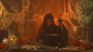 Siyah elbiseli bir cadı, kabaklarla, insan kafatası ve kemiklerle korkunç bir mutfak yaratıyor. Genç bir kadın ayin için haç alıyor. Cadılar Bayramı Konsepti