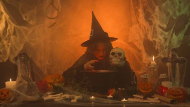 A witch in black robes conjures up a skull in a scary kitchen with pumpkins and potions. A young woman pulls a human skull from a cauldron and smiles frighteningly. Halloween Concept. clipart