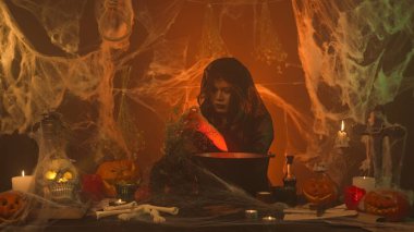 Siyah elbiseli cadı korkunç mutfakta balkabağı, insan kafatası ve kemiklerle iksir pişiriyor. Genç bir kadın tek tek kazan malzemesi ekliyor, ara sıra kameraya bakıyor. Cadılar Bayramı Konsepti.