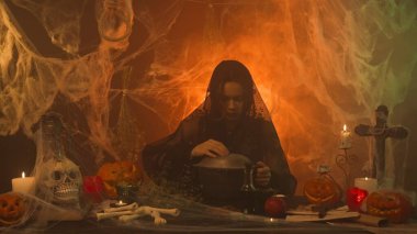 Siyah elbiseli bir cadı korkunç bir mutfakta balkabağı, insan kafatası ve kemiklerle iksir pişiriyor. Genç bir kadın bir kazanın kapağını açarak büyü yapıyor. Cadılar Bayramı Konsepti.