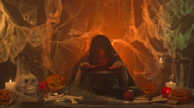 A witch in black clothing is cooking in cauldron in a scary kitchen with pumpkins and human skull and bones. A young woman speaks an incantation she adds ingredients to the potion. Halloween concept. clipart