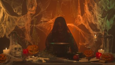 Siyah elbiseli bir cadı korkunç bir mutfakta kazanda yemek pişiriyor balkabakları, insan kafatası ve kemiklerle. Genç bir kadın sihirli bir söz söyler ve iksire malzemeler ekler. Cadılar Bayramı konsepti.