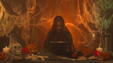 Siyah elbiseli bir cadı, ürkütücü bir mutfakta balkabaklarıyla, insan kafatası ve kemikleriyle oturan bir iksir üretir. Genç bir kadın sinsice gülümsüyor ve kazana bakıyor. Cadılar Bayramı Konsepti.