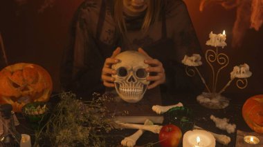 A witch in black clothing conjures up a skull sitting in a scary kitchen with pumpkins and potions. A young woman looks fearfully into the camera and touches a human skull. Halloween Concept. clipart