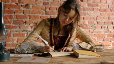 Ortaçağ dişisi eski bir tüy kalemle yazar. Rönesans elbisesi içinde antika genç bir kadın masada oturuyor ve eski parşömen kağıtlarına mürekkep sürülmüş eski kalem kalem ile mektup yazıyor..