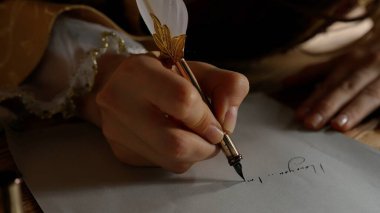 Ortaçağ dişisi eski bir tüy kalemle yazar. Rönesans elbisesi giymiş antika genç bir kadın masada oturuyor ve eski parşömen kâğıdına mürekkep sürülmüş eski kalem kalemle mektup yazıyor. Yakın çekim..
