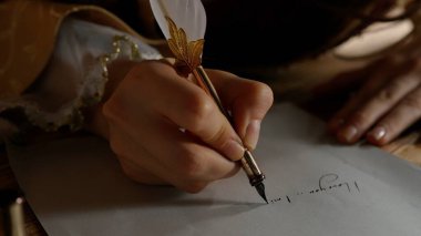 Ortaçağ dişisi eski bir tüy kalemle yazar. Rönesans elbisesi giymiş antika genç bir kadın masada oturuyor ve eski parşömen kâğıdına mürekkep sürülmüş eski kalem kalemle mektup yazıyor. Yakın çekim..