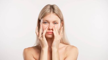 Çekici kadın model güzellik tedavisi yapıyor. Kameraya poz veren genç sarışın kadın beyaz stüdyo arka planında izole edilmiş parmaklarıyla pürüzsüz yüze dokunuyor. Güzel cilt bakımı konsepti