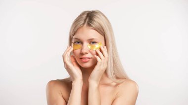 Çekici kadın model güzellik tedavisi yapıyor. Göz bandıyla kameraya poz veren genç sarışın kadın tenine dokunuyor, beyaz stüdyo arka planında izole edilmiş. Güzel cilt bakımı konsepti