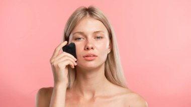 Çekici kadın model güzellik tedavisi yapıyor. Kameraya poz veren genç sarışın kadın, yüz hatlarına masaj yapan pembe stüdyo arka planında izole edilmiş bir kadın. Güzel cilt bakımı konsepti