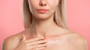 Çekici kadın model güzellik tedavisi yapıyor. Kameraya poz veren genç sarışın kadın köprücük kemiği bölgesine vücut losyonu sürüyor. Pembe stüdyo arka planında izole edilmiş. Yakın plan çekimi. Güzel cilt bakımı konsepti
