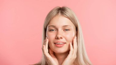 Çekici kadın model güzellik tedavisi yapıyor. Genç sarışın kadın kameraya poz veriyor, neşeli bir şekilde gülümsüyor ve tenine dokunuyor, pembe stüdyo arka planında soyutlanmış. Güzel cilt bakımı konsepti