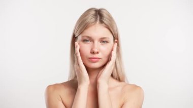 Çekici kadın model güzellik tedavisi yapıyor. Kameraya poz veren genç sarışın kadın beyaz stüdyo arka planında izole edilmiş parmaklarıyla pürüzsüz yüze dokunuyor. Güzel cilt bakımı konsepti