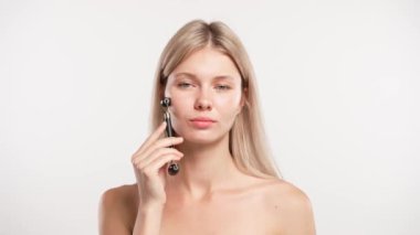 Çekici kadın model güzellik tedavisi yapıyor. Genç sarışın kadın kamera karşısında poz veriyor, yüz masajı yapıyor taş silindirli, beyaz stüdyo arka planında izole edilmiş. Güzel cilt bakımı konsepti