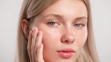 Çekici kadın model güzellik tedavisi yapıyor. Kameraya poz veren genç sarışın kadın, pürüzsüz yüz derisine dokunuyor, beyaz stüdyo arka planında izole edilmiş, yakın plan çekimi. Güzel cilt bakımı konsepti