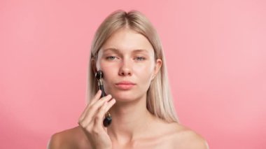 Çekici kadın model güzellik tedavisi yapıyor. Genç sarışın kadın kamera karşısında poz veriyor, yüz masajı yapıyor taş silindirli, pembe stüdyo arka planında izole edilmiş. Güzel cilt bakımı konsepti