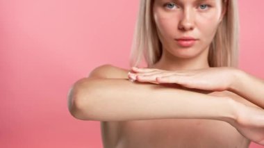Çekici kadın model güzellik tedavisi yapıyor. Kameraya poz veren genç sarışın kadın, koluna vücut kremi sürüyor, pembe stüdyo arka planında izole edilmiş, yakın çekim. Güzel cilt bakımı konsepti