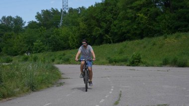 Bir adam, güneşli bir günde, uzun ağaçlar ve yeşilliklerle çevrili, asfalt bir yolda bisiklet sürüyor..