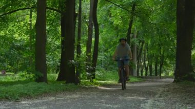 Bir adam, güneşli bir günde, uzun ağaçlar ve yeşilliklerle çevrili, asfalt bir yolda bisiklet sürüyor..