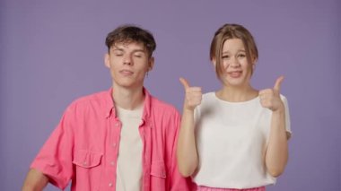 Stüdyoda poz veren genç adam ve kadın. Mor arka planda izole edilmiş pembe beyaz elbiseli kız ve erkek arkadaş pozitifler. Kız baş parmağını kaldırıyor, erkek el işareti gösteriyor..