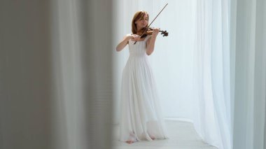 Beyaz akışkan elbiseli bir kadın parlak minimalist bir odada keman çalıyor ve beyaz perdeler sallanıyor. Huzurlu bir atmosfer ve büyüleyici bir kemancının müzik performansı..