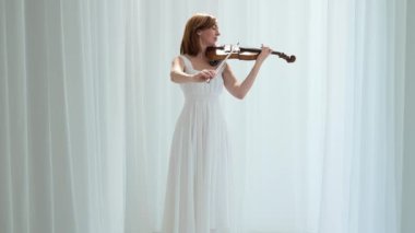 Beyaz akışkan elbiseli bir kadın parlak minimalist bir odada keman çalıyor ve beyaz perdeler sallanıyor. Huzurlu bir atmosfer ve büyüleyici bir kemancının müzik performansı..
