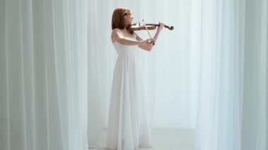 Beyaz akışkan elbiseli bir kadın parlak minimalist bir odada keman çalıyor ve beyaz perdeler sallanıyor. Huzurlu bir atmosfer ve büyüleyici bir kemancının müzik performansı..