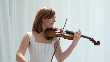 Beyaz akışkan elbiseli bir kadın parlak minimalist bir odada keman çalıyor ve beyaz perdeler sallanıyor. Huzurlu bir atmosfer ve büyüleyici bir kemancının müzik performansı..