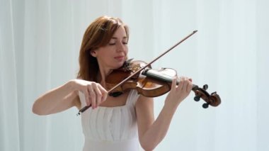 Beyaz akışkan elbiseli bir kadın parlak minimalist bir odada keman çalıyor ve beyaz perdeler sallanıyor. Huzurlu bir atmosfer ve büyüleyici bir kemancının müzik performansı..