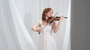 Beyaz akışkan elbiseli bir kadın parlak minimalist bir odada keman çalıyor ve beyaz perdeler sallanıyor. Huzurlu bir atmosfer ve büyüleyici bir kemancının müzik performansı..