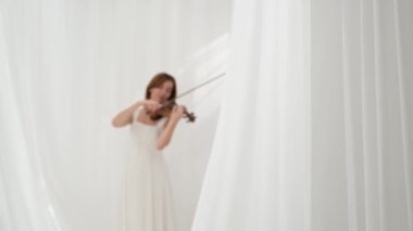 Beyaz akışkan elbiseli bir kadın parlak minimalist bir odada keman çalıyor ve beyaz perdeler sallanıyor. Huzurlu bir atmosfer ve büyüleyici bir kemancının müzik performansı..
