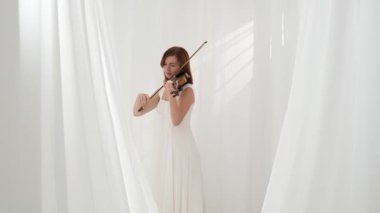 Beyaz akışkan elbiseli bir kadın parlak minimalist bir odada keman çalıyor ve beyaz perdeler sallanıyor. Huzurlu bir atmosfer ve büyüleyici bir kemancının müzik performansı..