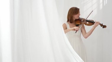 Beyaz akışkan elbiseli bir kadın parlak minimalist bir odada keman çalıyor ve beyaz perdeler sallanıyor. Huzurlu bir atmosfer ve büyüleyici bir kemancının müzik performansı..