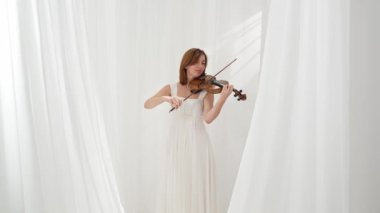 Beyaz akışkan elbiseli bir kadın parlak minimalist bir odada keman çalıyor ve beyaz perdeler sallanıyor. Huzurlu bir atmosfer ve büyüleyici bir kemancının müzik performansı..