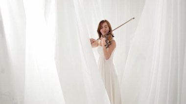 Beyaz akışkan elbiseli bir kadın parlak minimalist bir odada keman çalıyor ve beyaz perdeler sallanıyor. Huzurlu bir atmosfer ve büyüleyici bir kemancının müzik performansı..
