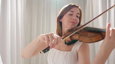 Beyaz akışkan elbiseli bir kadın parlak minimalist bir odada keman çalıyor ve beyaz perdeler sallanıyor. Huzurlu bir atmosfer ve büyüleyici bir kemancının müzik performansı..