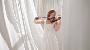 Beyaz akışkan elbiseli bir kadın parlak minimalist bir odada keman çalıyor ve beyaz perdeler sallanıyor. Huzurlu bir atmosfer ve büyüleyici bir kemancının müzik performansı..