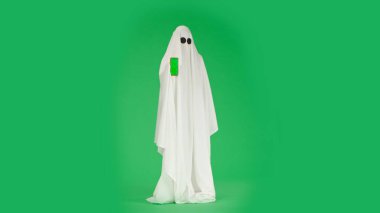 Kameraya poz veren hayalet gibi giyinmiş biri. Beyaz çarşaflı cadılar bayramı hayaleti kameraya akıllı telefon modelini gösteriyor, krom anahtar yeşil ekranda izole edilmiş, tam boy. Ürkütücü konsept.