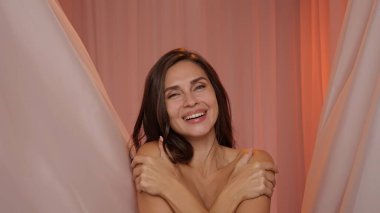 Stüdyoda poz veren güzel bir kadın model. Makyajlı ve saç stili olan kadın kamera tebessümlerine, boyun ve göğüs bölgesine, uçan pembe kumaşlar stüdyo arka planına bakıyor. Güzellik ve sağlık konsepti.